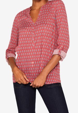 Mujer lleva una blusa roja de manga larga con estampado, puños remangados y vaqueros azul oscuro, manos juntadas delante de la cintura.