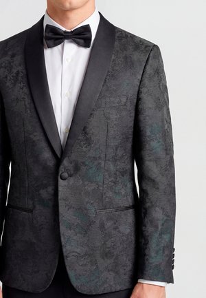 Hombre vistiendo una chaqueta de esmoquin negra de jacquard floral con solapas tipo chal de satén, camisa blanca de vestir y pajarita negra, visible desde el cuello hasta la cadera.