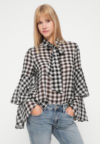 Melnbalts raibs blūze ar viļņotām piedurknēm un saistītu kakla daļu, izgatavota no viegla, caurspīdīga auduma. Kombinēta ar augstu jostasvietu biksēm.