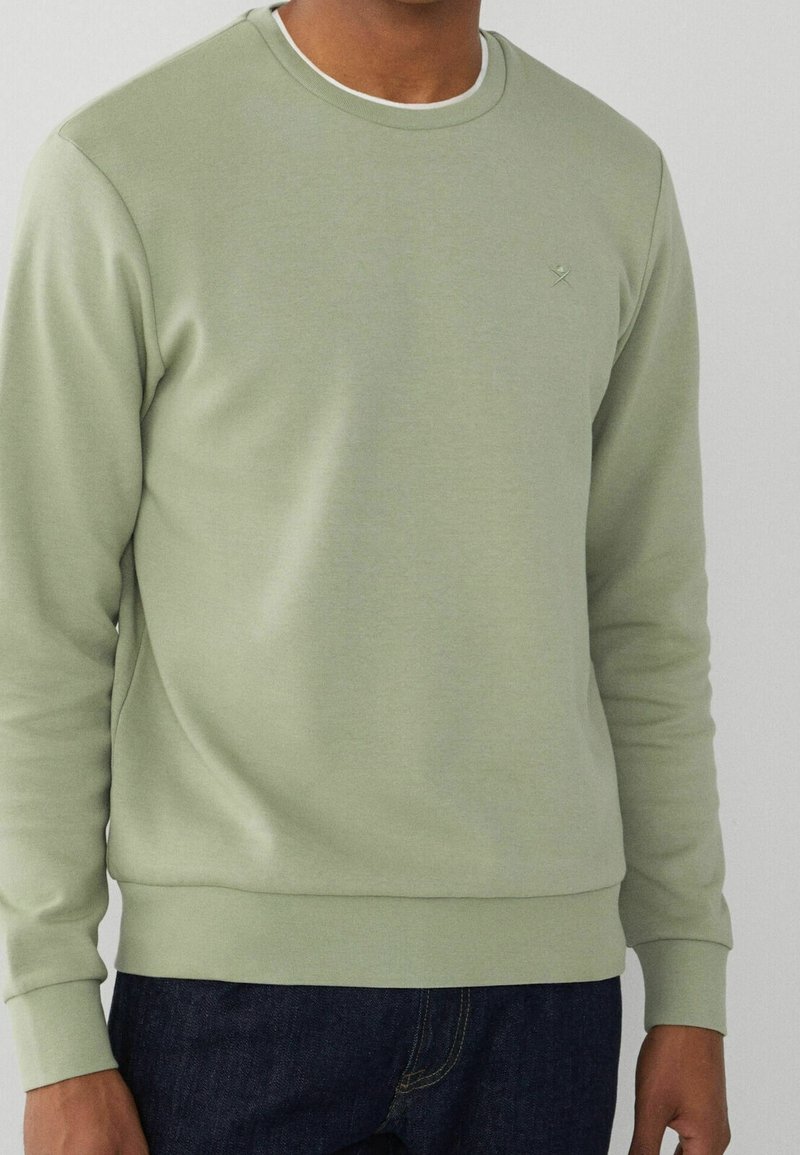 Homme portant un sweat-shirt vert clair à col rond avec des manches longues et un jean bleu foncé devant un fond uni.