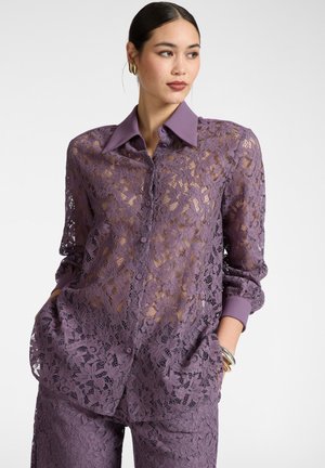 Mujer con cabello oscuro recogido en un moño bajo, lleva una blusa morada de encaje floral con botones y pantalones a juego, complementados con pendientes de aro dorados y pulseras.