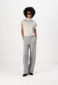 Haut beige à manches courtes avec un col montant, associé à un pantalon large gris à motifs, accessoirisé avec des lunettes de soleil noires et des chaussures à talons.