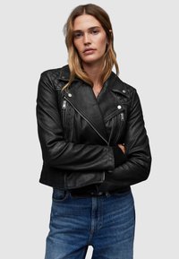 Giubbotto da biker in pelle nera con zip e bottoni a pressione argentati; design accorciato, spalle trapuntate, indossato su un capo nero, abbinato a jeans blu.