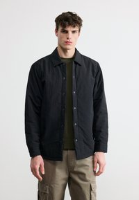 Νεαρός άνδρας με κοντά σγουρά μαλλιά, φοράει ένα μαύρο σακάκι με κουμπιά, φανελένιο πουκάμισο σε ελιά πράσινο χρώμα και μπεζ παντελόνι cargo, μπροστά σε λευκό φόντο.