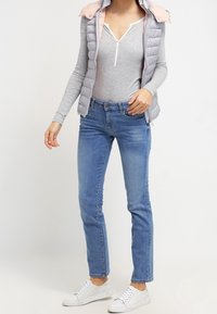 Kvinna som bär en ljusgrå långärmad tröja, en ärmlös rosa och grå puffväst, blå jeans och vita sneakers, stående mot vit bakgrund.