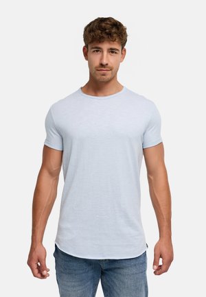 Man met kort krullend haar, gekleed in een lichtblauw T-shirt met korte mouwen en een blauwe spijkerbroek, staand tegen een effen witte achtergrond.