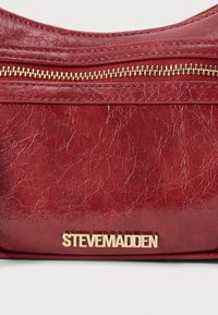 Punane tekstureeritud nahk kott, millel on kuldne tõmblukk ja kuldne "Steve Madden" brändi logo.