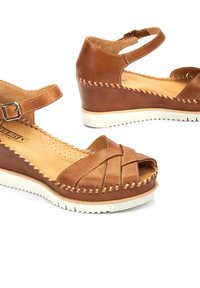 Sandales compensées en cuir marron avec un design entrecroisé, bout ouvert, sangle de cheville ajustable et semelle en caoutchouc blanc avec bord texturé.