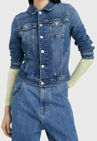 Denim jacket featuring a collar, front pockets, and silver buttons. Worn over a patterned green long-sleeve shirt, with a fitted design.