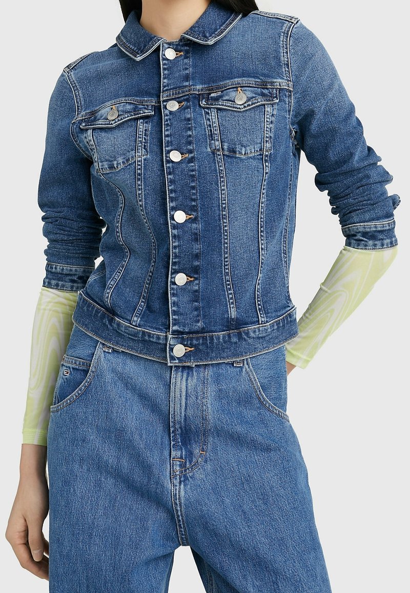 Denim jacket featuring a collar, front pockets, and silver buttons. Worn over a patterned green long-sleeve shirt, with a fitted design.