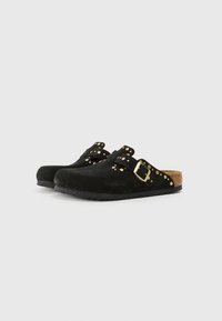 Sabots en suede noir avec une forme arrondie, semelle en liège et accents en clous dorés le long de la sangle, dotés d'une fermeture à boucle sur le côté.