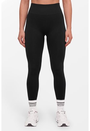 Persoon draagt zwarte leggings met hoge taille, witte enkelsokken met zwarte strepen en tekst, en witte sportschoenen en staat rechtop.