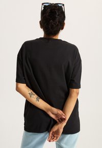 Camiseta de algodón negra con mangas cortas, corte relajado. Presenta un escote redondo y se combina con jeans de un azul claro. Tatuaje intrincado visible en el brazo.