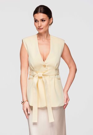 Kobieta z ciemnymi włosami związanymi do tyłu, ubrana w bezrękawnik w kolorze kremowym z głębokim dekoltem w kształcie litery V, z paskiem w talii oraz jasnobeżową spódnicę, pozująca na jednolitym tle.