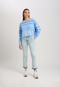 Hellblau gestreifter Pullover mit lockerer Passform, Kordelzug-Saum, kombiniert mit hellen Jeans und weißen High-Top-Sneakers.