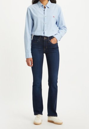 Personne portant une chemise bleu clair à boutons, un jean slim bleu foncé et des baskets blanches, debout avec une main dans la poche.