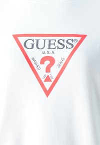 Biela tričko s červeným trojuholníkovým logom s nápismi "GUESS" a "U.S.A." v čiernej farbe. Obsahuje texty "WASHED" a "JEANS" spolu s grafikou otáznik.
