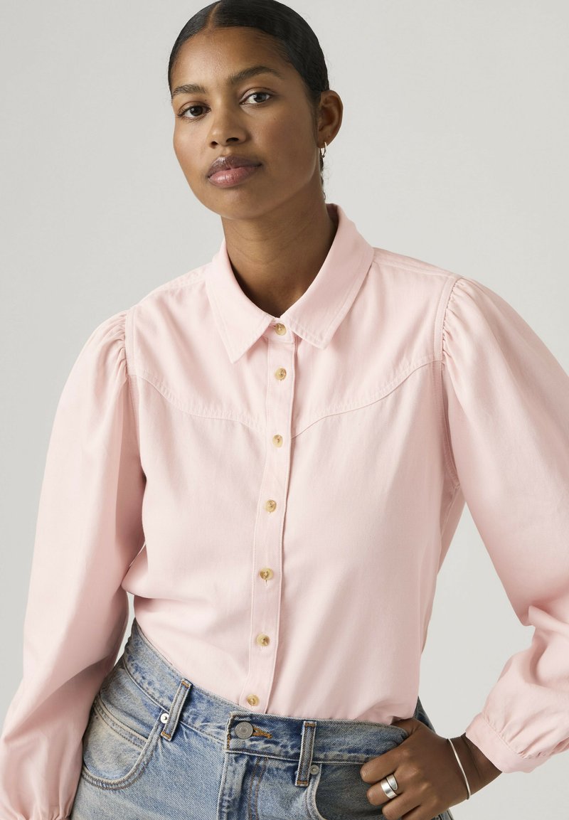 Femme aux cheveux foncés tirés en arrière, portant une chemise rose clair boutonnée avec des manches ballons et un jean bleu clair, posant avec une main sur la hanche.