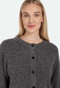 Šedý vlněný cardigan s kulatým výstřihem, černými knoflíky a texturovaným pleteným vzorem, který má volný střih a dlouhé rukávy.