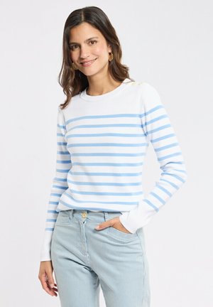 Femme aux cheveux bruns portant une chemise blanche à manches longues avec des rayures horizontales bleu clair et un pantalon rayé bleu clair, souriante avec une main dans la poche.