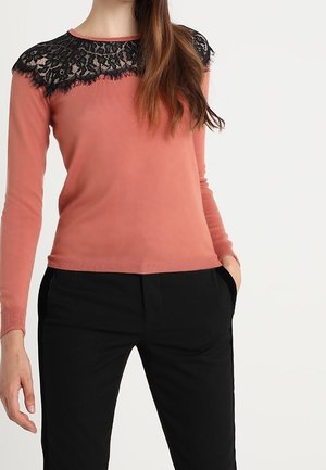 Pull à manches longues en corail doux avec un bodice en dentelle noire. Il présente un col rond et des poignets côtelés. Porté avec un pantalon noir.
