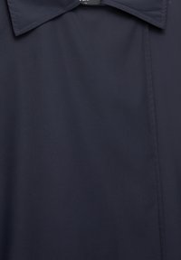 Chemise bleu marine foncé avec col, coutures visibles et texture de tissu lisse, suspendue à plat sur un fond uni.