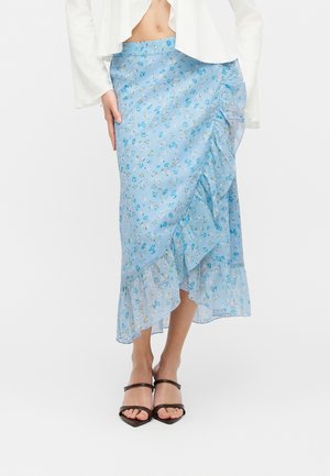 Žena oblečená v svetlomodrej midi sukni s kvetinovým vzorom a volánikovým lemom, doplnená čiernymi sandálmi s otvorenou špičkou a bielou skrátenou blúzkou.