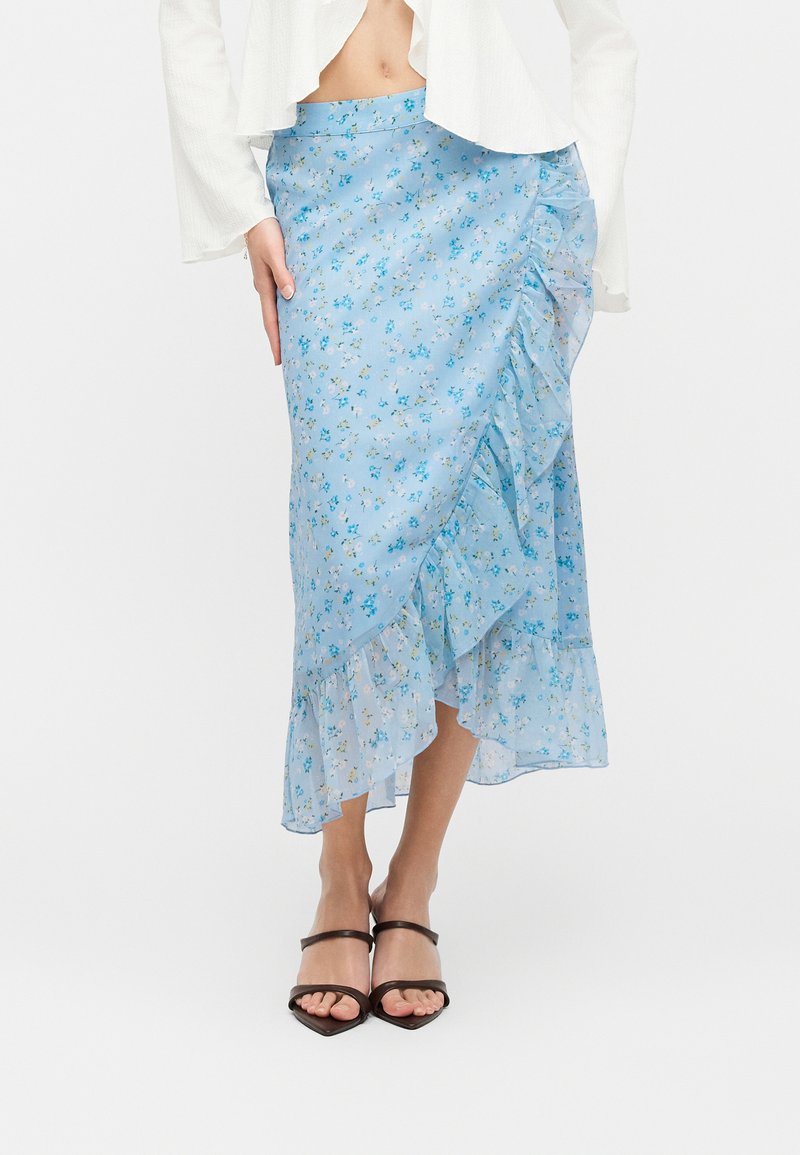 Γυναίκα που φοράει φούστα μέσης μήκους με ανοιχτό μπλε λουλουδάτο σχέδιο και βολάν στο τελείωμα, συνδυασμένη με μαύρες ανοιχτές γόβες και άσπρο κοντό μπλουζάκι.