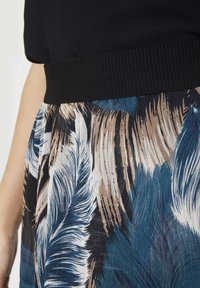Haut noir avec large ceinture côtelée associé à une jupe arborant un grand motif de plumes bleues, blanches et brunes sur un tissu texturé.