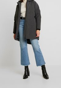 Chaqueta acolchada larga gris con cierre de cremallera y capucha, combinada con pantalones vaqueros claros de campana con dobladillos deshilachados y botines negros de tacón.