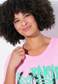 Roze T-shirt met ronde hals, met groen graffiti-achtige tekst en patronen. Zachte stof met een licht gestructureerde uitstraling. Close-up weergave.