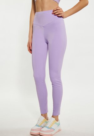Donna che indossa leggings lilla a vita alta e scarpe da ginnastica bianche e colorate, in piedi con una mano sul fianco su sfondo bianco.