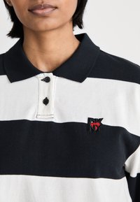 Persona vistiendo una camisa polo de rayas horizontales negras y blancas con un pequeño logotipo de corazón rojo y las iniciales negras DKNY en el pecho.