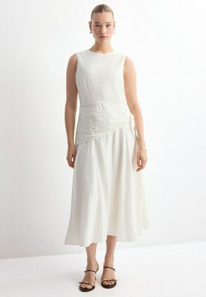 Frau steht und trägt ein ärmelloses weißes Kleid mit Spitzen-Detail an der Taille und schwarze offene Sandaletten mit Absatz vor einem schlichten weißen Hintergrund.