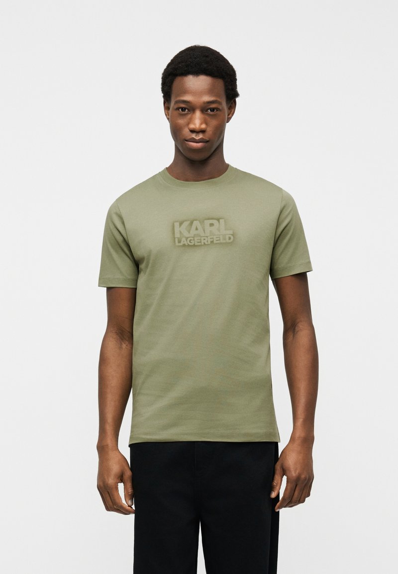 Jeune homme portant un t-shirt vert olive avec le texte "KARL LAGERFELD" sur la poitrine et un pantalon noir, posé devant un fond uni.