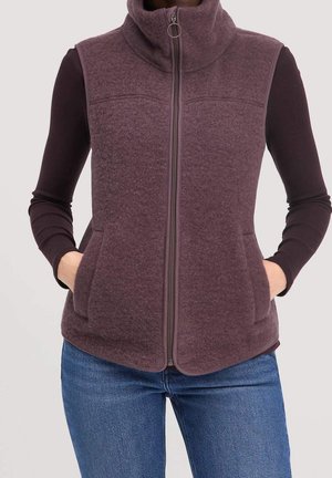 Bezrękawnik z jasnofioletowego polaru zapinany na zamek, noszony na czarnej bluzce z długim rękawem, z rękami w przednich kieszeniach, w połączeniu z niebieskimi dżinsami.