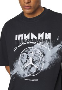Close-up of person wearing black t-shirt with white "JORDAN" text, basketball graphic, and Jumpman logo over a splattered paint design.