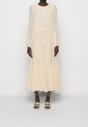 Kvinde iført en tynd, cremefarvet maxikjole med lange ærmer, snøre i taljen og lagdelt kant, kombineret med hvide sandaler med stropper.