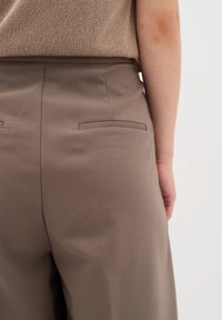 Gros plan d'une personne portant un pantalon marron taille haute avec des poches passepoilées dans le dos et un haut en maille beige rentré à l'intérieur.