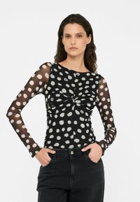 Top nero con maniche lunghe in rete trasparente, caratterizzato da pois bianchi, design davanti attorcigliato e una silhouette aderente. Abbinato a jeans neri.