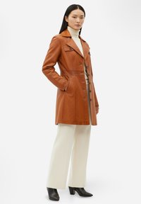 Brun läderjacka med skräddarsydd design, hakskärning och knappstängning. Bärs över en krämfärgad polotröja och vita vida byxor.