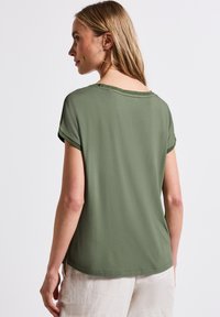 Olivgrön t-shirt med kort ärm och avslappnad passform, rundad halsringning och upprullade ärmslut; slät textur och subtila sömmar på baksidan.