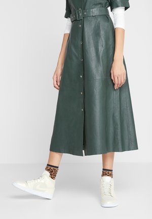 Femme portant une robe midi en cuir vert foncé avec ceinture, des baskets montantes blanches et des chaussettes courtes à imprimé léopard, debout avec un pied légèrement levé.
