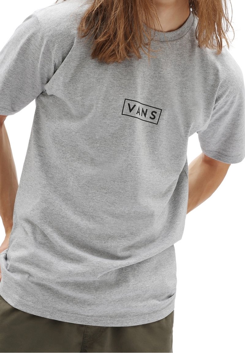 Personne portant un t-shirt Vans gris à manches courtes et un pantalon vert olive, les mains posées derrière le dos.