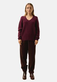 Bordowy sweter w serek o teksturowanym splocie, w połączeniu z brązowymi spodniami z чудя. Model nosi ciemno-brązowe botki za kostkę.