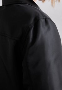 Zwarte jas met gladde, glanzende stof, een gestructureerde kraag en een getailleerde vorm, met een close-up van de schoudernaden.