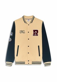 Veste varsity beige et noire avec col, poignets et ourlet rayés, ornée de patches et du texte "Redskins Div. 1984 Team Leader."