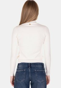 Donna con capelli lunghi e lisci che indossa una maglia bianca aderente a maniche lunghe e jeans di jeans blu, vista da dietro su uno sfondo semplice.