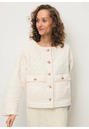 Kobieta o kręconych włosach, ubrana w jasną pikowaną kurtkę z guzikami i dwoma dużymi przednimi kieszeniami, patrząca w prawo.