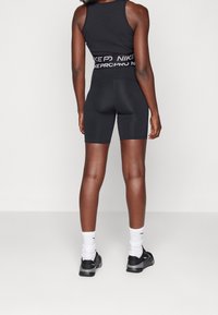 Osoba ubrana w czarne szorty i top Nike Pro, białe skarpetki i czarne sneakersy, stojąca tyłem na jednolitym tle.